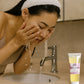 Woman washing her face with water in a bathroom, with a tube of skincare product in the foreground.