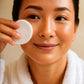 Woman applying a facial product with a cotton pad on a neutral background